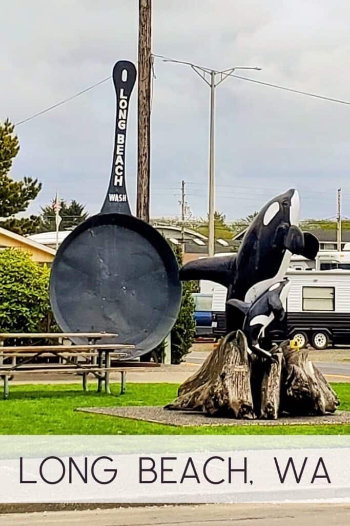 A roadside scene in Long Beach, Washington featuring the "World's Largest Frying Pan" sculpture—a tall, black metal skillet standing upright. Next to it is a wood-carved sculpture of two orca whales jumping out of a stump. In the background are a green lawn, picnic tables, and a white travel trailer under a cloudy sky.