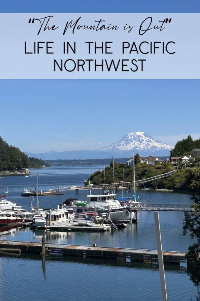 Life in the Pacific Northwest A bright, sunny day at a Pacific Northwest marina with Mount Rainier towering in the background. Several white boats are docked in the calm blue water of the harbor, surrounded by lush green trees and coastal homes. A clear, blue sky sits above the snow-capped volcanic peak. A blue text overlay at the top reads: "'The Mountain is Out' LIFE IN THE PACIFIC NORTHWEST."