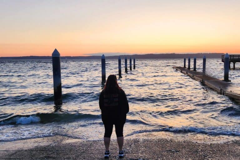 A woman looking out over the water as the sun sets over the mountains in the distance.