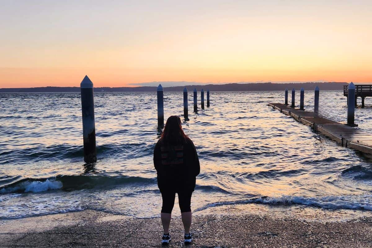 A woman looking out over the water as the sun sets over the mountains in the distance.