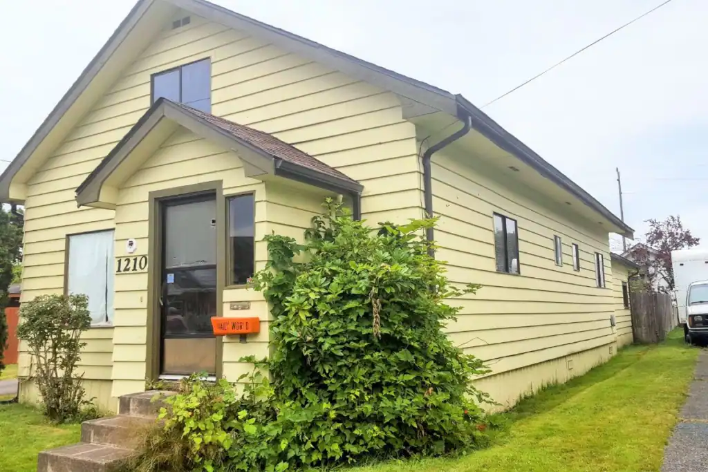 Kurt Cobain Childhood Home 2018 The exterior of Kurt Cobain's childhood home in 2018, showing pale yellow green siding an orange Daily World mailbox and overgrown bushes before restoration.