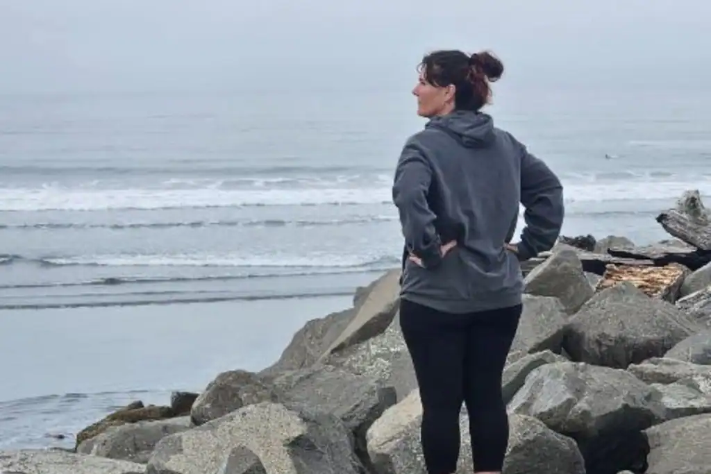 Washington Coast Solo Midlife Travel A woman in a grey hoodie looking out at the ocean waves from a rocky jetty on a cloudy day along the Washington coast.