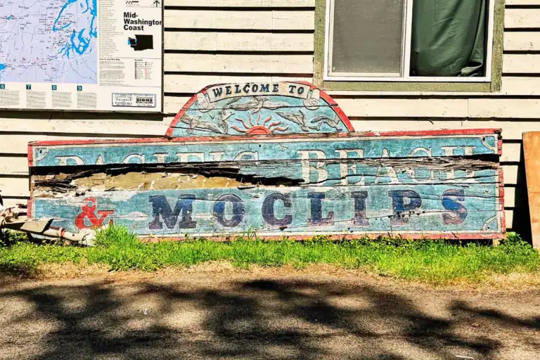 A weathered, vintage wooden welcome sign that reads 'Welcome to Pacific Beach & Moclips' with cracking paint next to a map of the mid-Washington coast.
