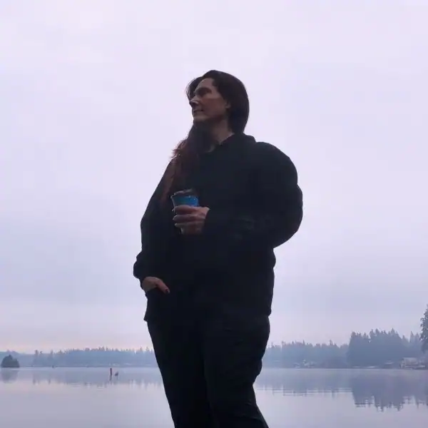 PNW Travel Girl blogger holding a coffee cup and looking out over a misty Pacific Northwest lake and treeline during a solo travel trip.
