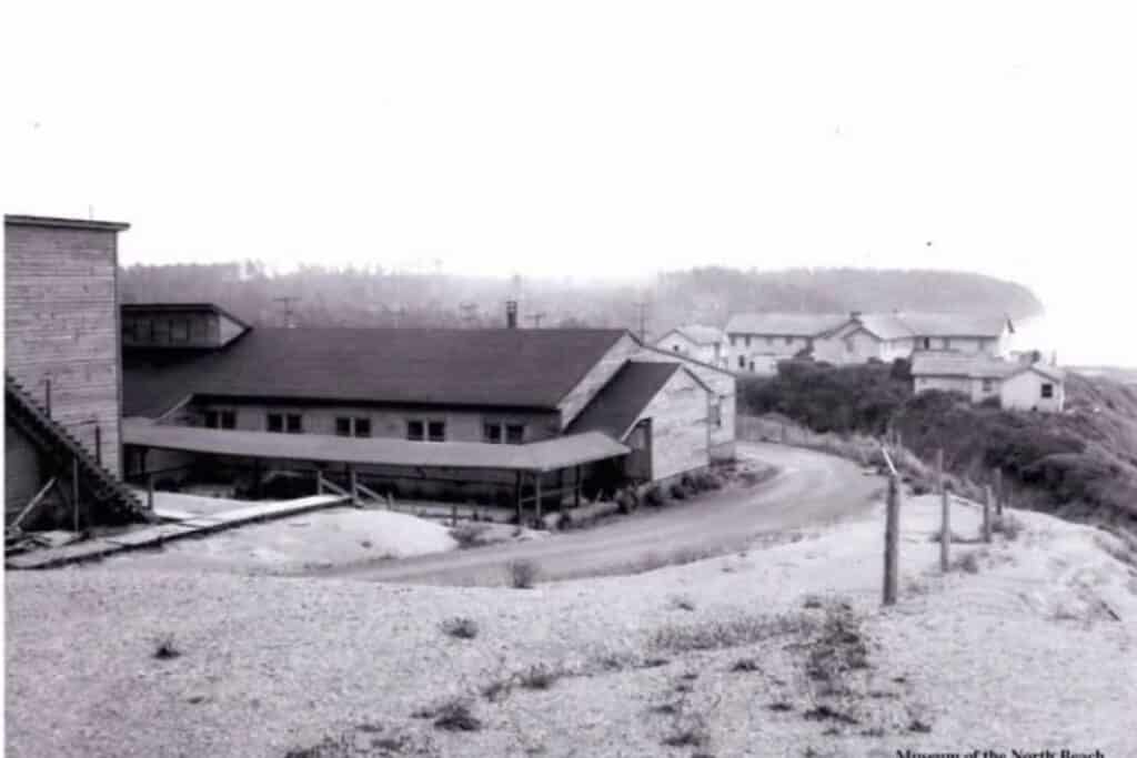 Historic photo of the newly designated NAVFAC Pacific Beach installation in 1958, courtesy of the Museum of the North Beach. Historical black and white photograph from July 1958 showing the Navy Base at Pacific Beach Washington with wooden military buildings, a winding gravel access road, fences, and the old Pacific Beach Hotel visible on the ocean bluff.