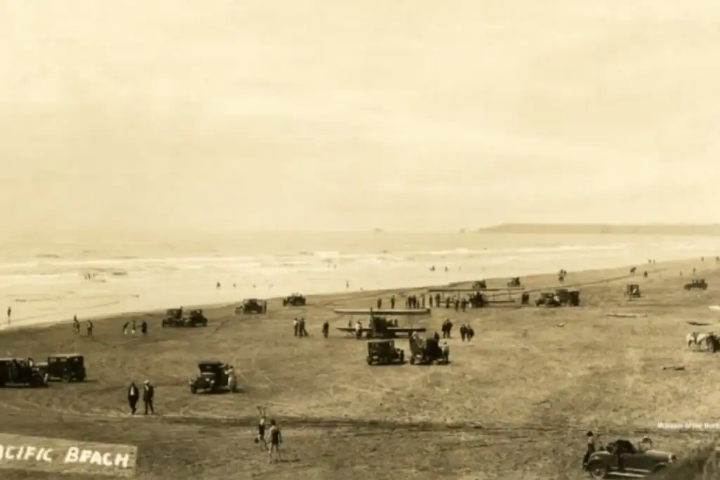 Vintage photo of early tourists, cars, and an airplane parked on the sand at Pacific Beach, courtesy of the Museum of the North Beach. Historical sepia photograph from the Museum of the North Beach showing an expansive Pacific Beach shoreline packed with 1920s vintage automobiles and a biplane parked directly on the hard sand near crowds of people.