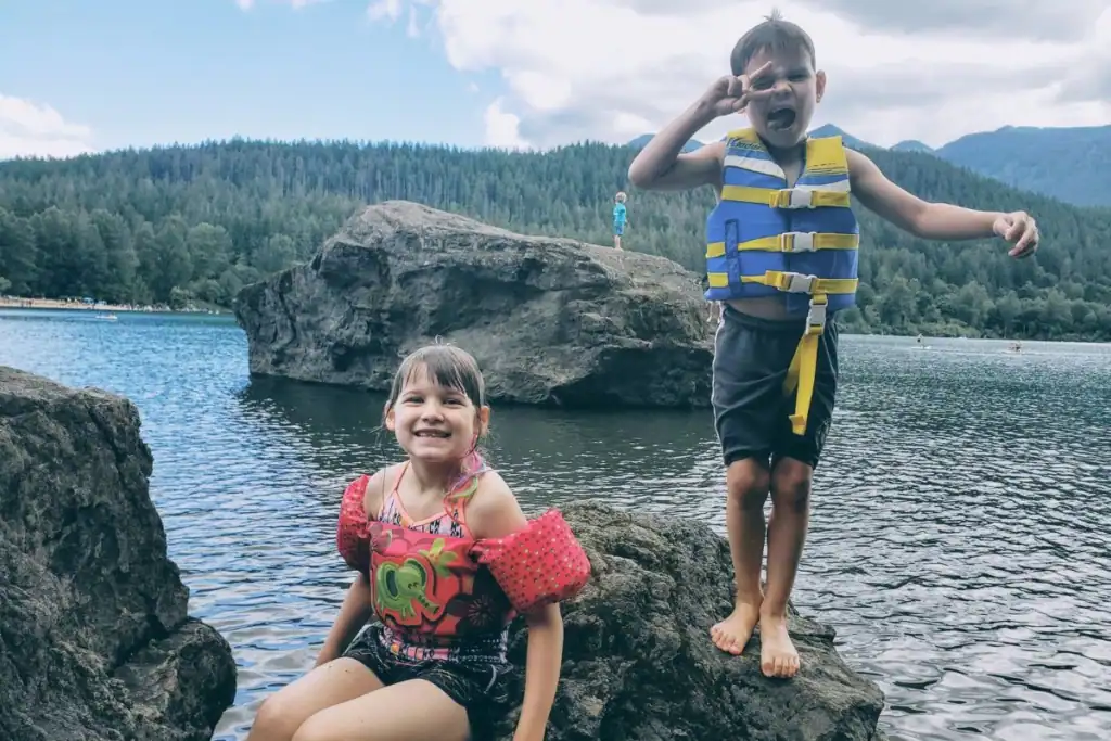 Two smiling children wearing swim gear sit and stand on shoreline rocks beside Rattlesnake Lake, Washington, with forested mountains in the background.