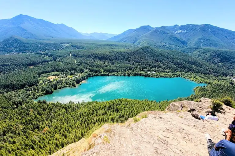 Panoramic view of turquoise Rattlesnake Lake surrounded by evergreen forests and Cascade Mountains from the rocky Rattlesnake Ledge viewpoint near North Bend, Washington.