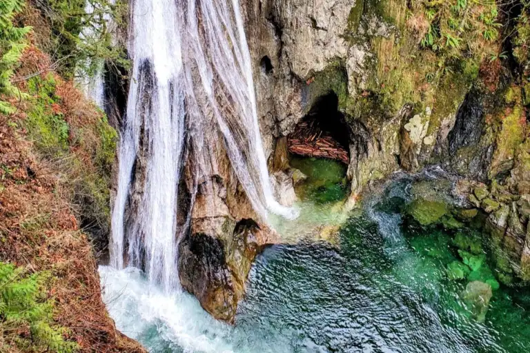 A close-up view of a rushing waterfall cascading into a crystal-clear turquoise river pool surrounded by mossy rocks and a small cave alcove along the Twin Falls Trail in North Bend Washington.