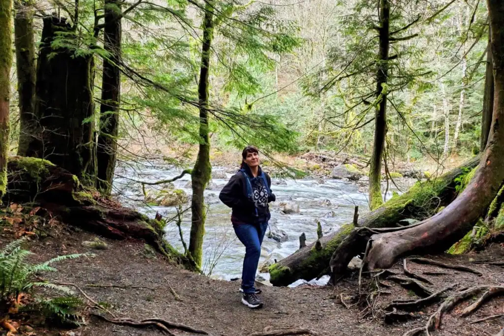 Woman standing on a forest trail beside the rocky South Fork Snoqualmie River, surrounded by moss-covered trees, exposed roots, and ferns near Twin Falls in Washington.
