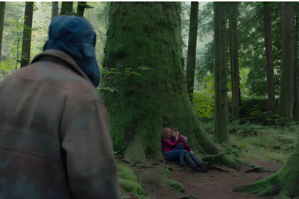 Scene from Twin Peaks showing a person in a blue hooded jacket approaching a large moss-covered tree, with two people sitting at its base in a dense evergreen forest.