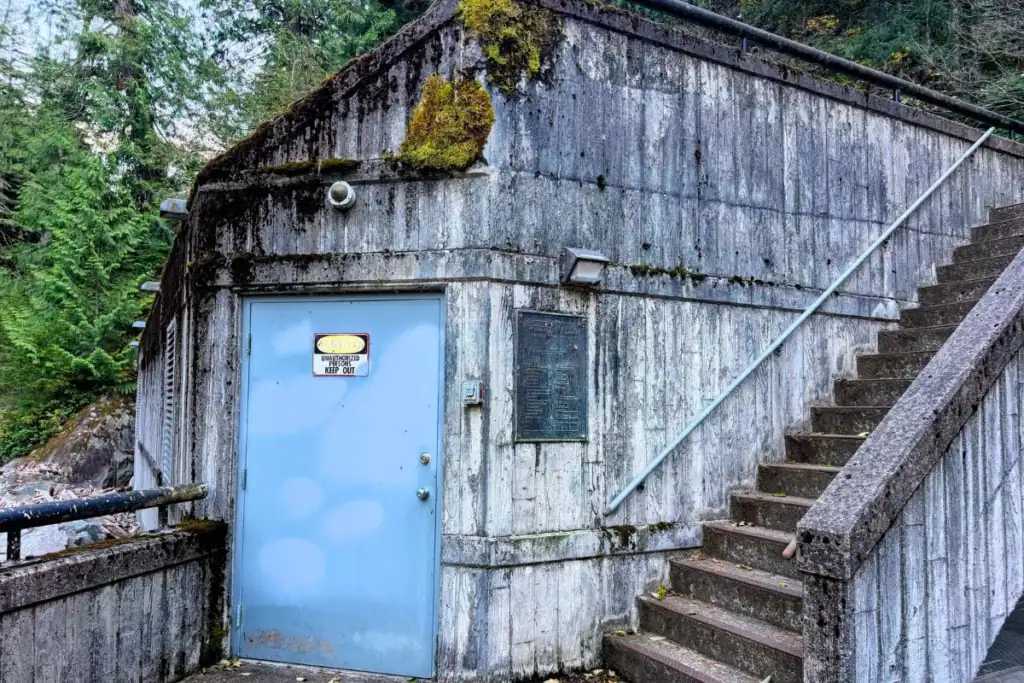 Weathered concrete hydroelectric facility building with a blue metal door, exterior staircase, and moss-covered walls near Weeks Falls at Olallie State Park.